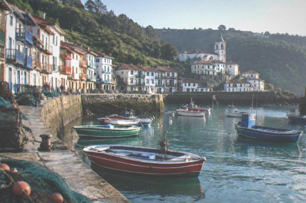 A white coastal village with a church tower above a small harbour and green slopes