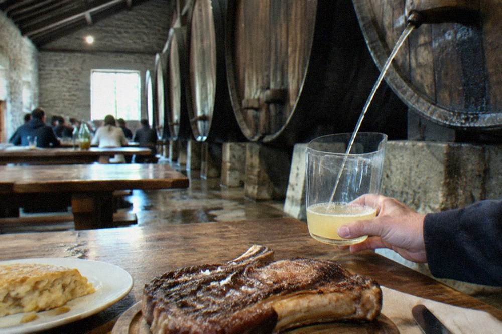 A glass of pale Basque cider held up in a dimly lit cider cellar