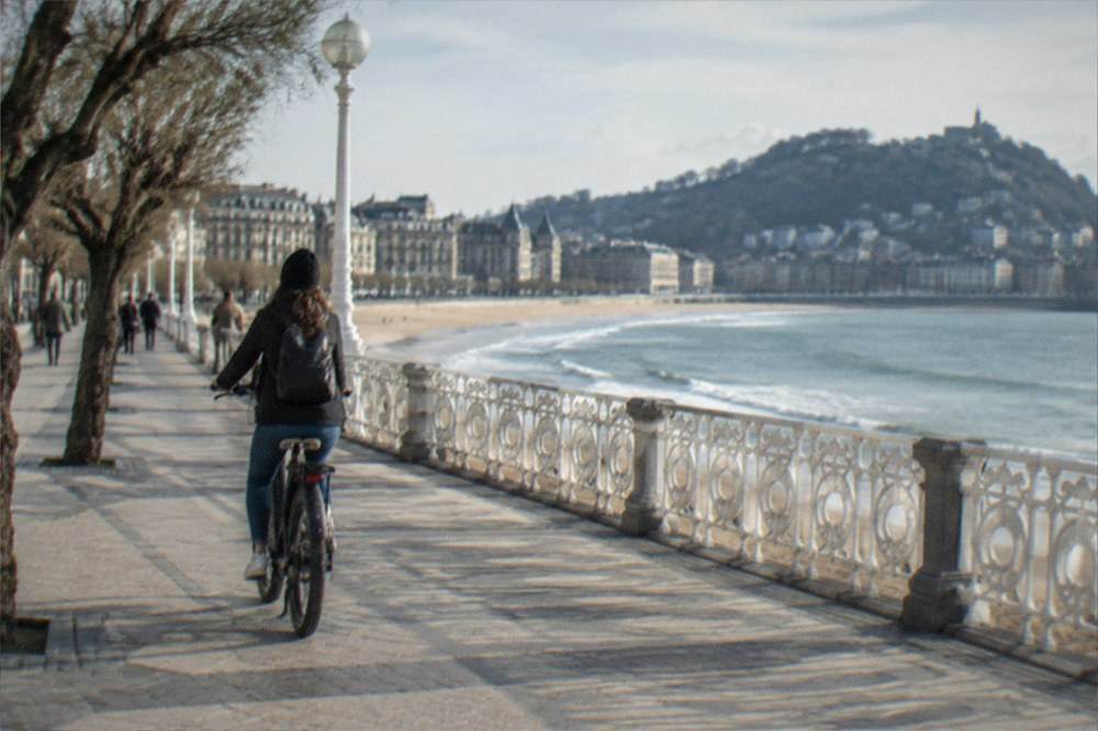 A cyclist riding an electric bike along a wide seafront promenade in San Sebastián