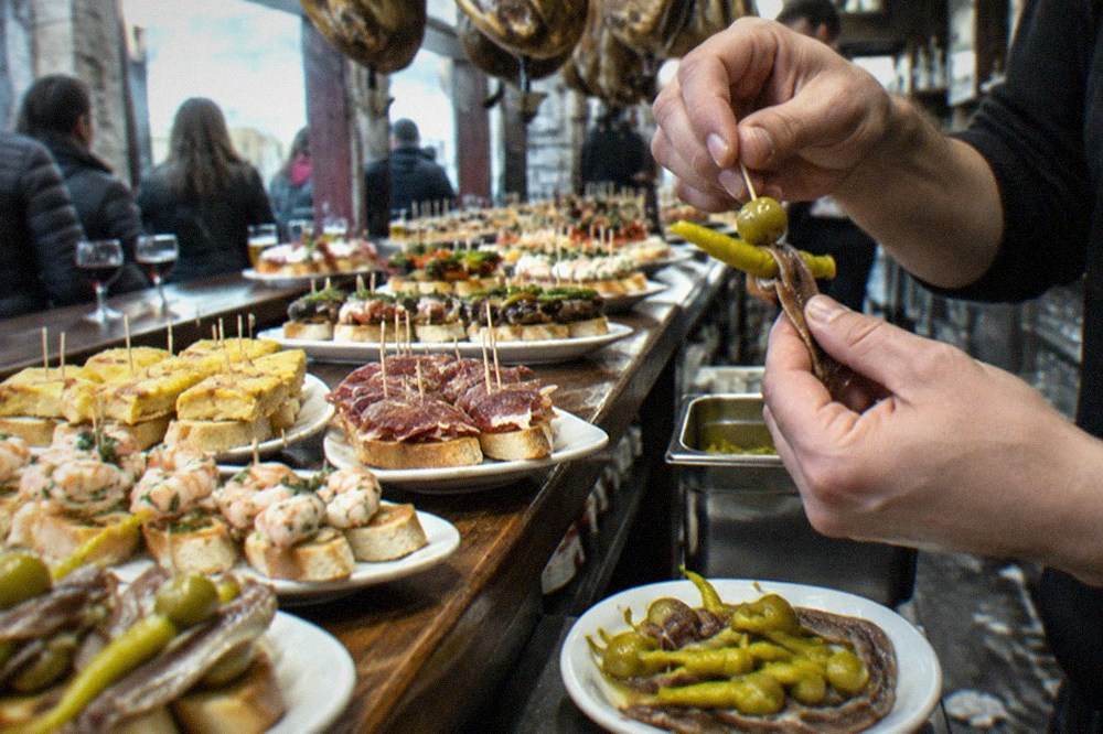 Hands assembling gilda pintxos on a bar counter, threading an anchovy, a pickled chilli and a green olive onto a toothpick