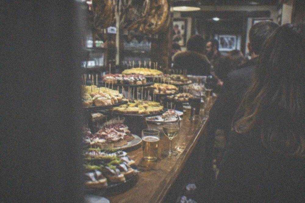 A crowded pintxo bar counter in the San Sebastián Old Town at evening, with skewers and small drinks on the counter
