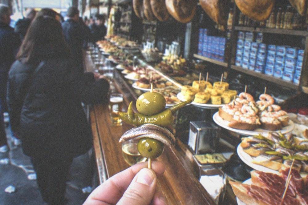 A gilda pintxo held up in front of a pintxo bar shelf stacked with anchovy tins and jars of pickled chillis