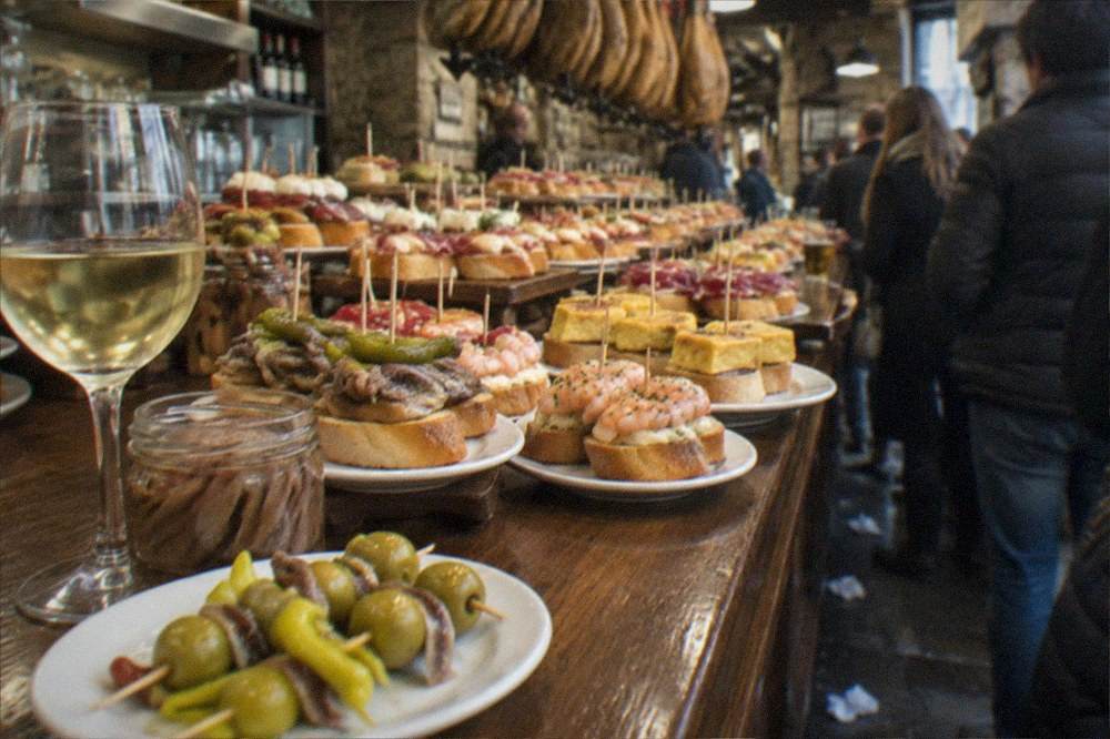 Home-made gilda pintxos on a small plate with a jar of anchovies and a glass of white wine on a kitchen table