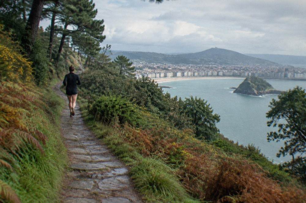 A stone path climbing a wooded hill above a bay with a city below