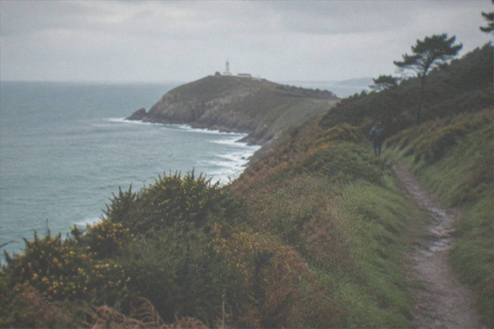 A grassy coastal headland path with the sea on one side and a lighthouse in the distance