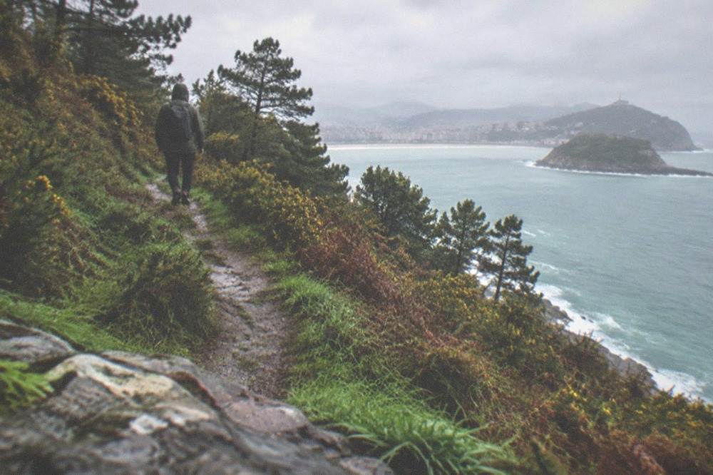 A hiker in a rain jacket walking a green ridge path above the sea