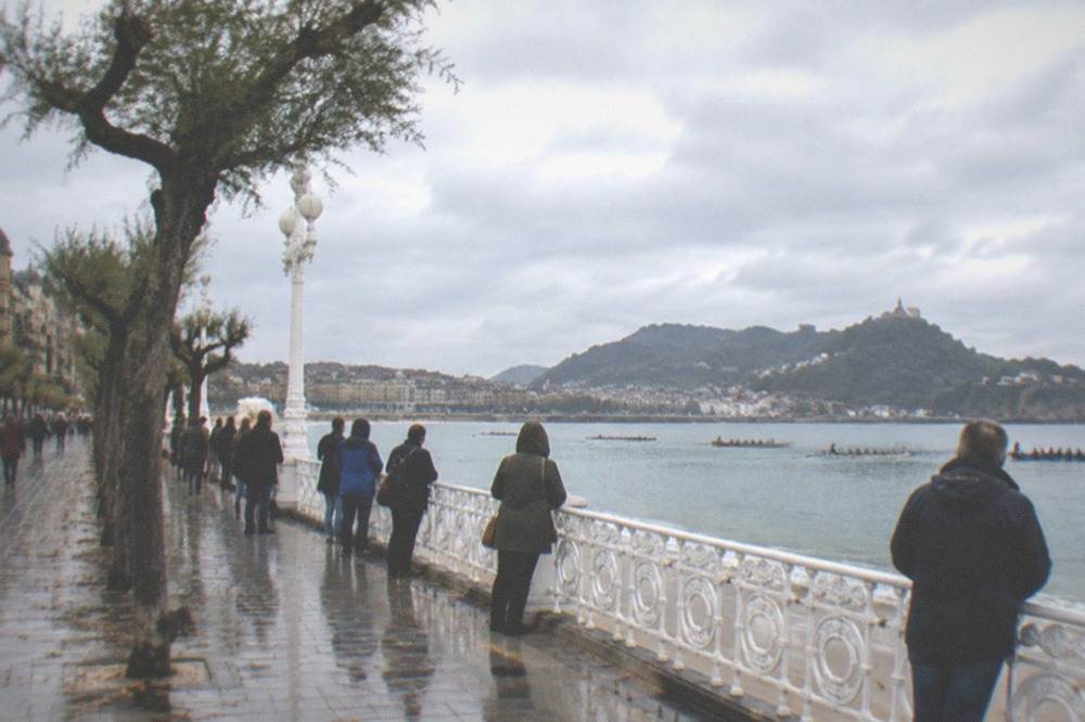 Spectators in rain jackets watching traineras from the promenade with grey sky over the bay