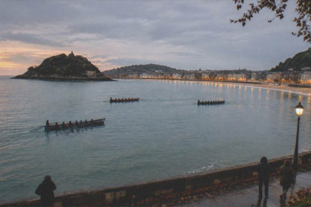 Traineras on the water at sunset with the illuminated bayfront of San Sebastián behind them