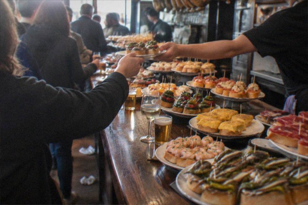 A bartender handing a small plate of pintxos to a customer at a busy bar counter in San Sebastián