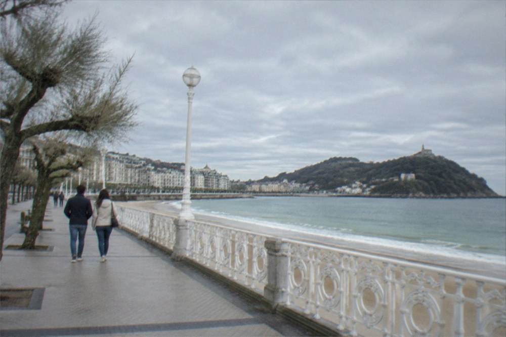 A couple in light jackets walking the La Concha promenade under a partly cloudy sky