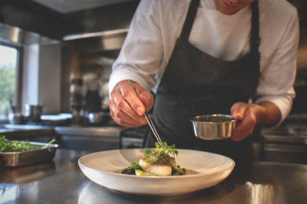 A chef plating a dish in a professional kitchen with stainless steel counters