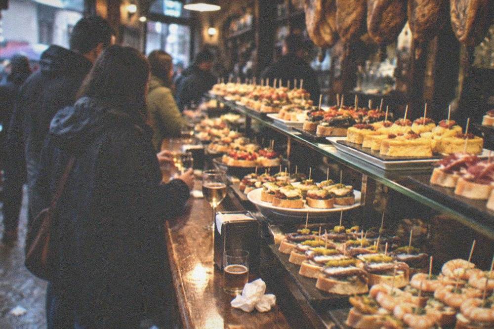 Rows of pintxos arranged on a bar counter under the lights of a Basque tavern