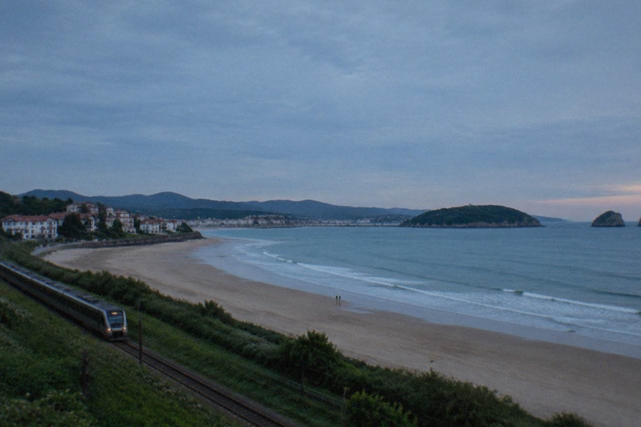 A train travelling along a green coastline toward a bay with a sandy beach and an island
