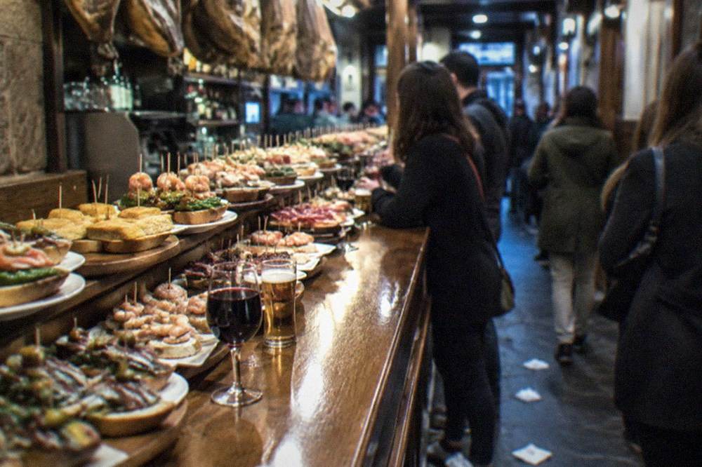 Friends standing at a pintxo bar counter with small glasses of wine in the old town of San Sebastián