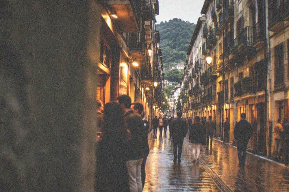 Narrow old town street in San Sebastián at dusk with lit bar fronts and people walking between them