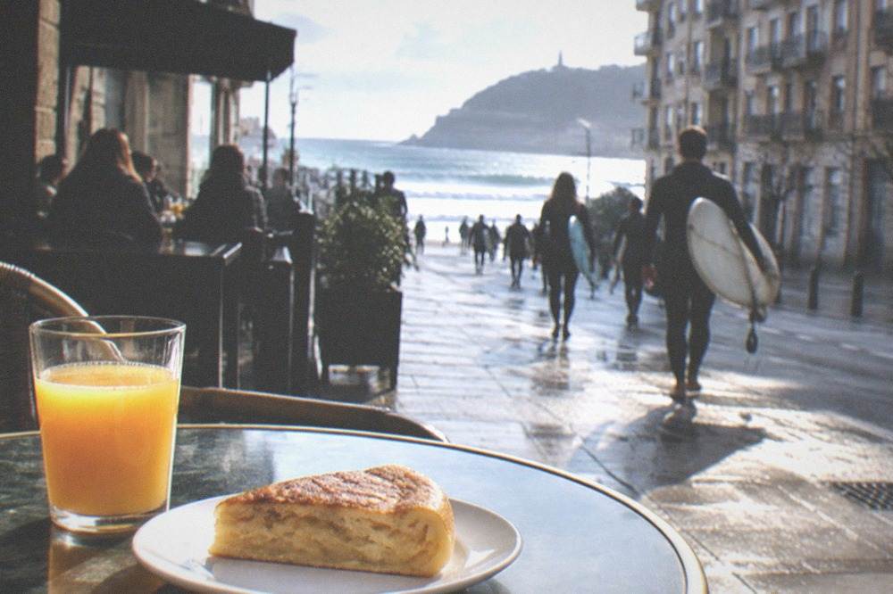 A slice of potato tortilla on a small plate beside a glass of orange juice on a café table