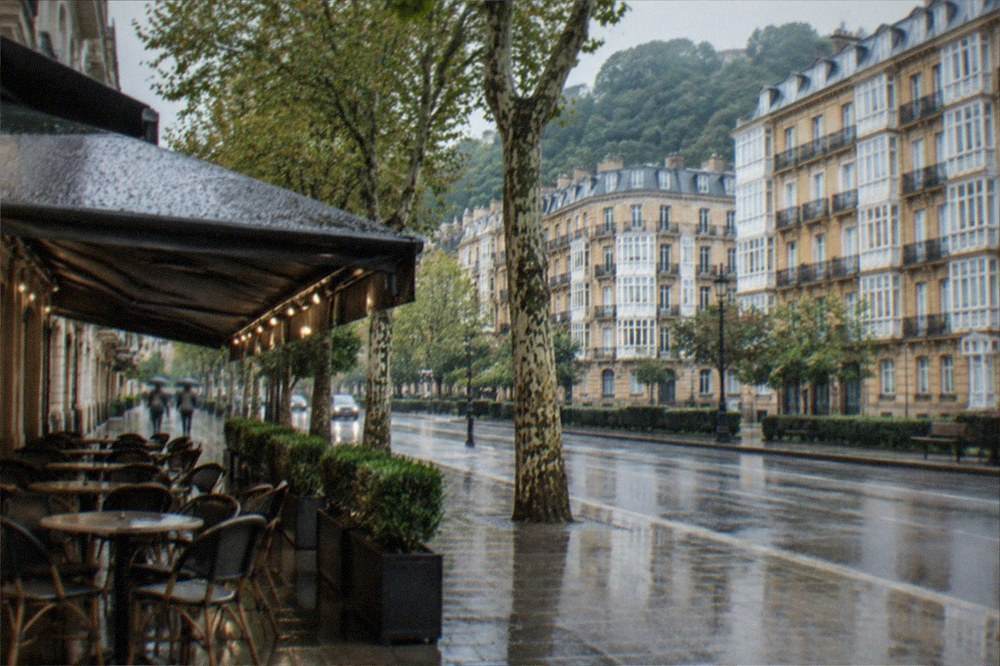 A covered café terrace with empty chairs under light rain beside a wet street in San Sebastián