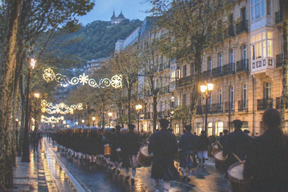 Rows of drummers and barrel carriers parading at night through a decorated street in San Sebastián