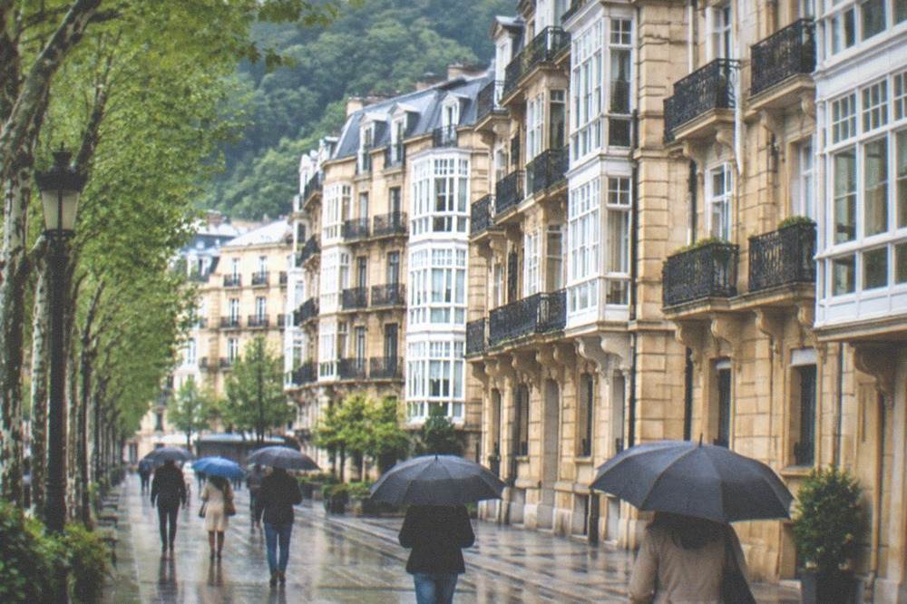 People walking with umbrellas past elegant Belle Epoque buildings on a wet street in central San Sebastián