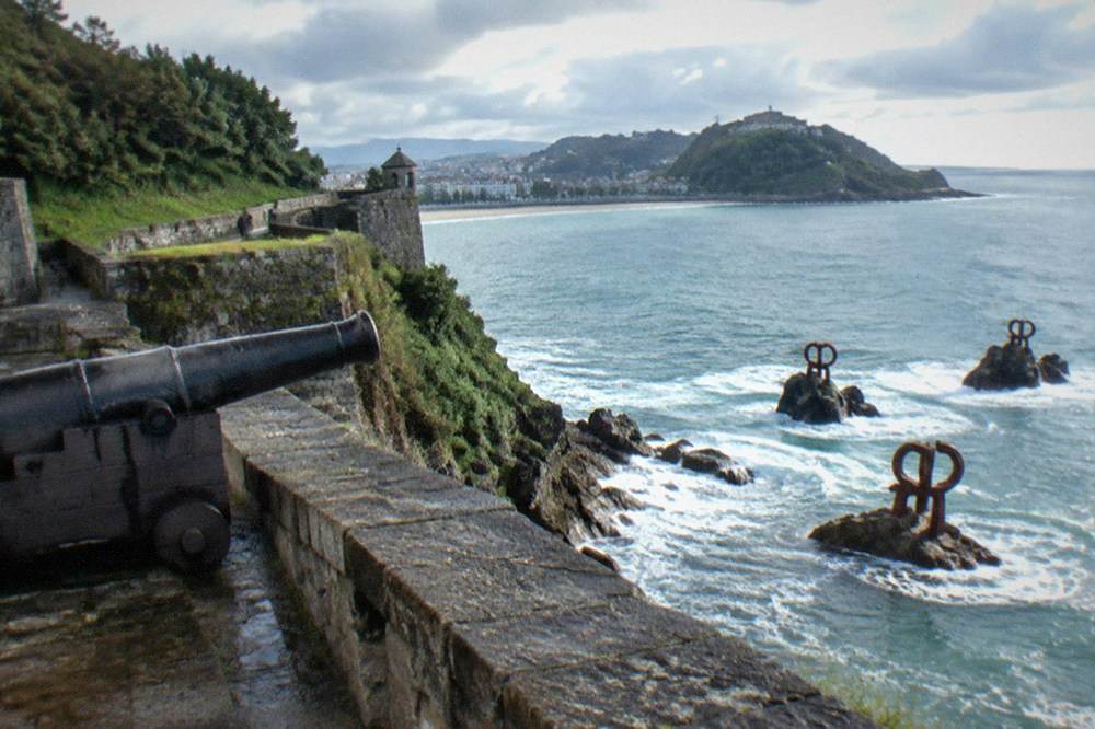 Three large steel sculptures standing on rocks at the water's edge, with waves breaking around them
