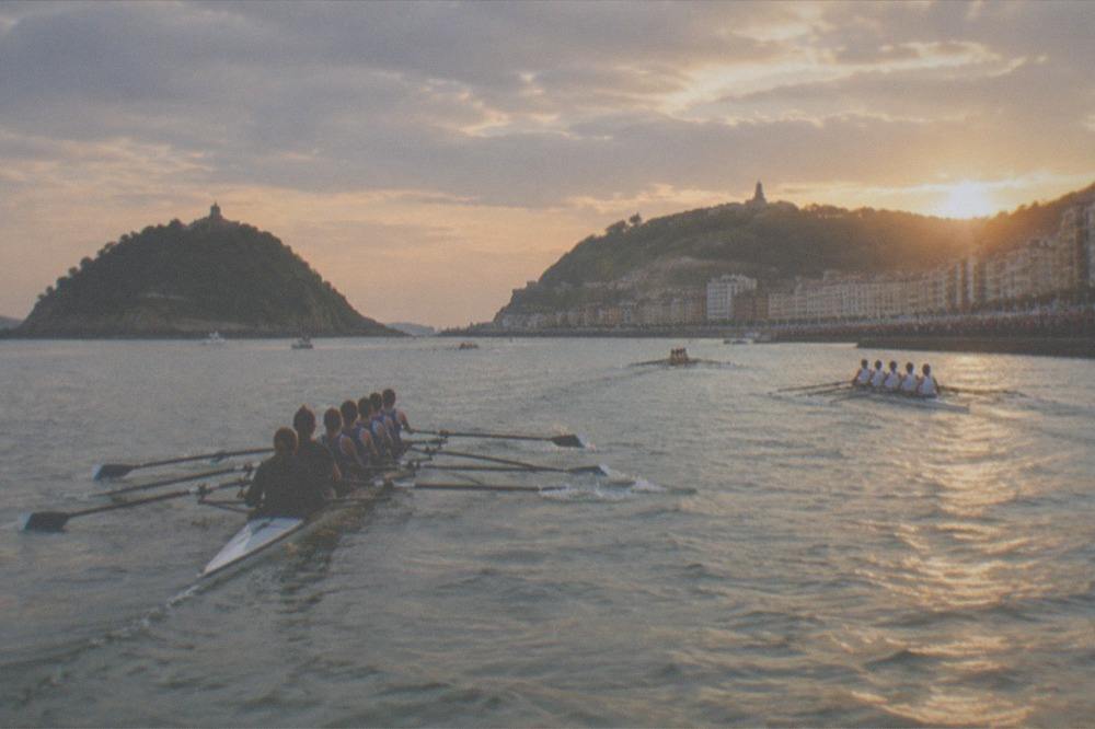 Rowing boats racing in a bay in front of a crowded seafront, with spectators along the railing