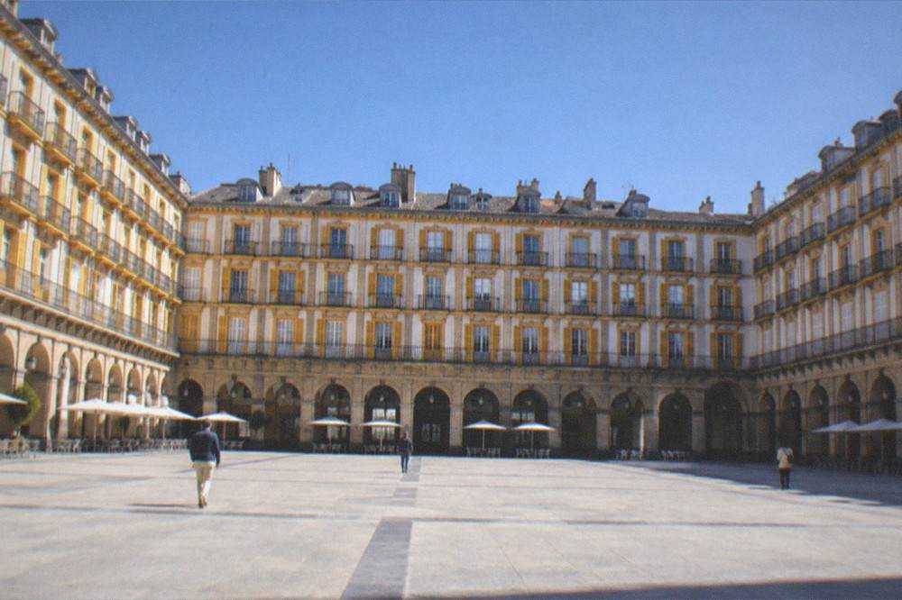 A wide plaza with tall buildings and numbered balconies around a colonnade, empty in morning light