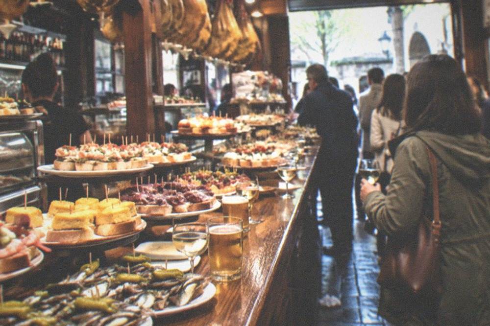 A pintxo bar counter in the Old Town of San Sebastián crowded with small plates and drinks