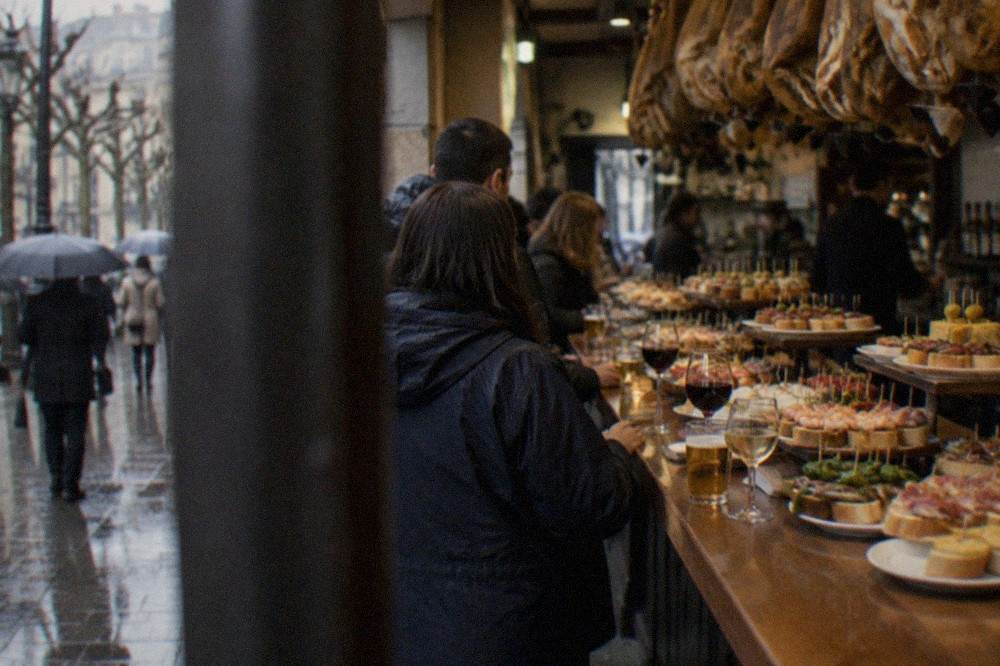 A busy pintxo bar counter in San Sebastián with trays of small skewered snacks and glasses of wine