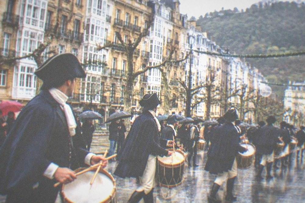 Rows of drummers in 19th-century costume marching through a decorated street in the city centre
