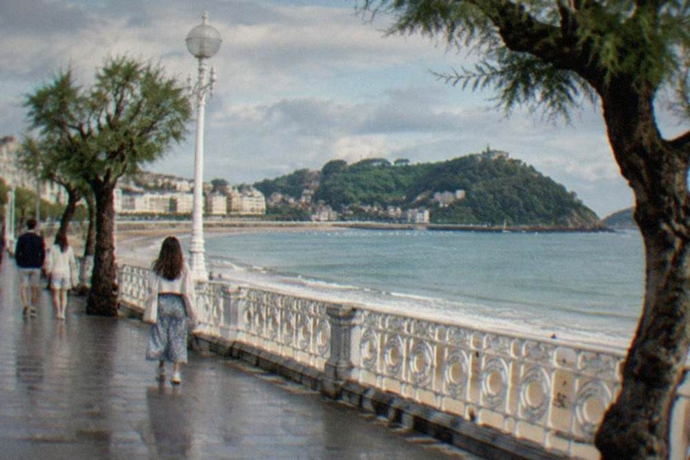 People walking along the La Concha promenade in San Sebastián under a summer sky with white railings and the bay beyond