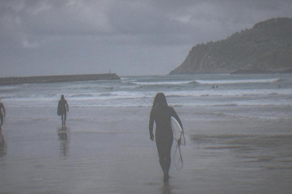 Surfers in wetsuits walking across an empty Zurriola beach under grey skies