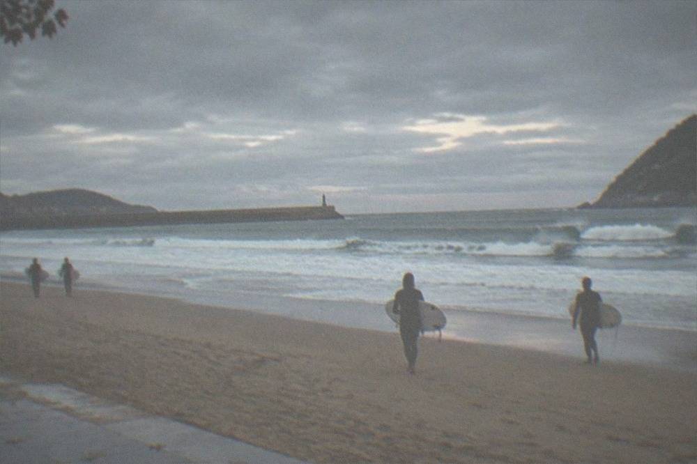 Surfers in wetsuits walking across Zurriola beach in San Sebastián under heavy grey clouds