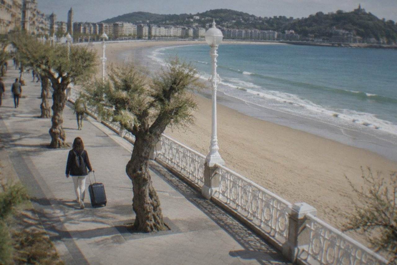 A traveller pulling a suitcase along a tree-lined promenade beside a wide sandy bay
