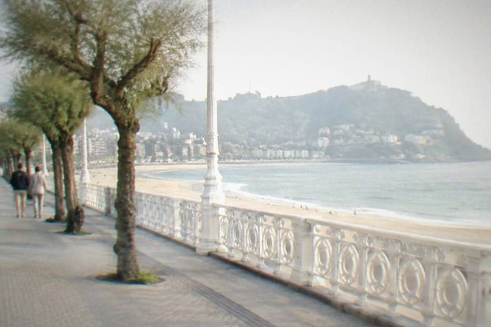 A seafront promenade with a railing, a crescent beach and hills beyond in late afternoon light