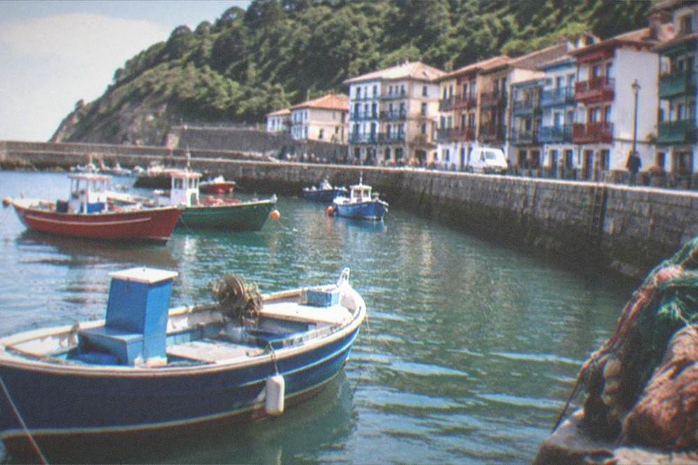 A coastal road view with a small boat in a harbour and shop-lined quayside buildings