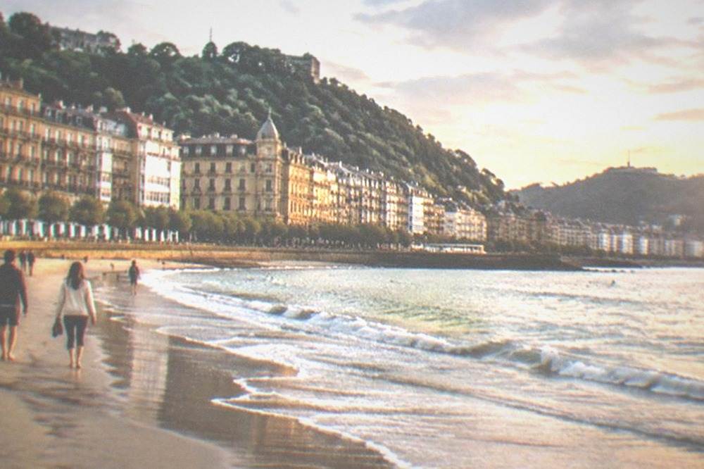 People walking on a wide sandy beach with a bay and hills behind
