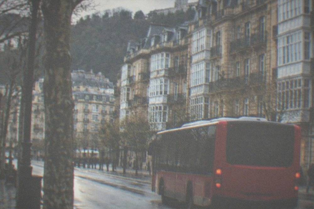 A red city bus pulling away from a stop on a tree-lined San Sebastián street