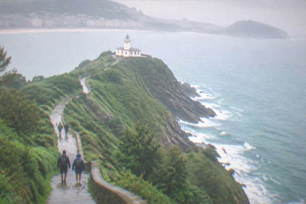 A coastal path climbing green cliffs above the sea toward the Ulia lighthouse