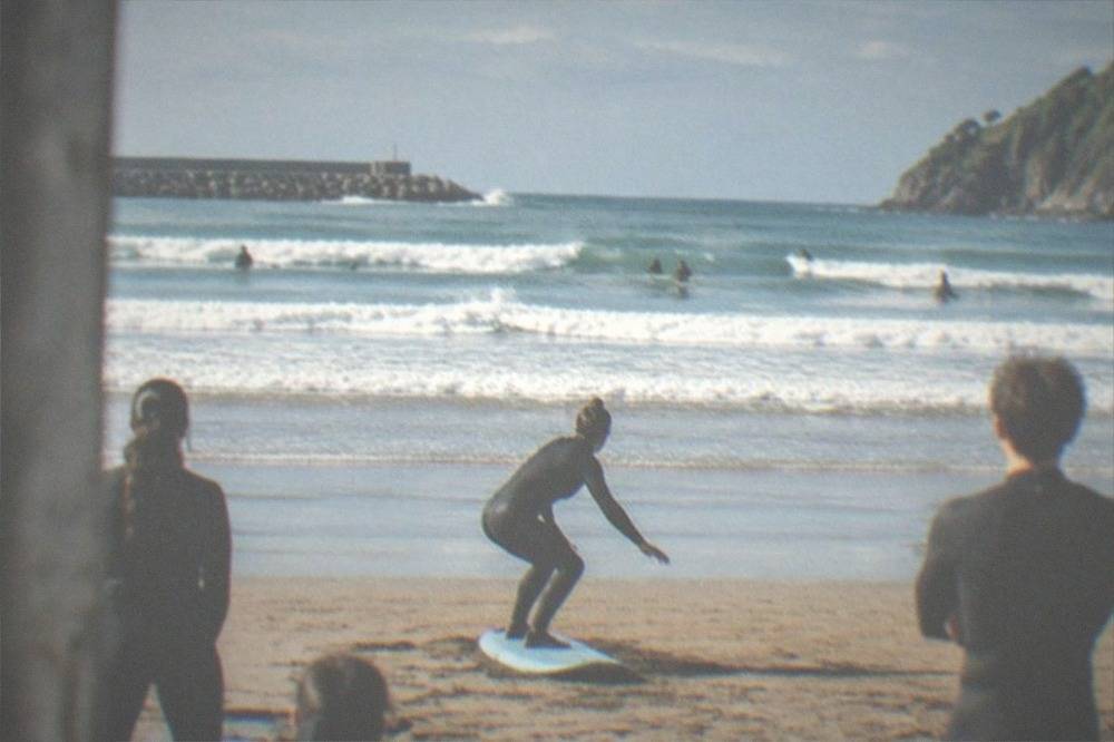 A surf instructor demonstrating a pop-up stance on the sand at Zurriola beach to a small group of learners