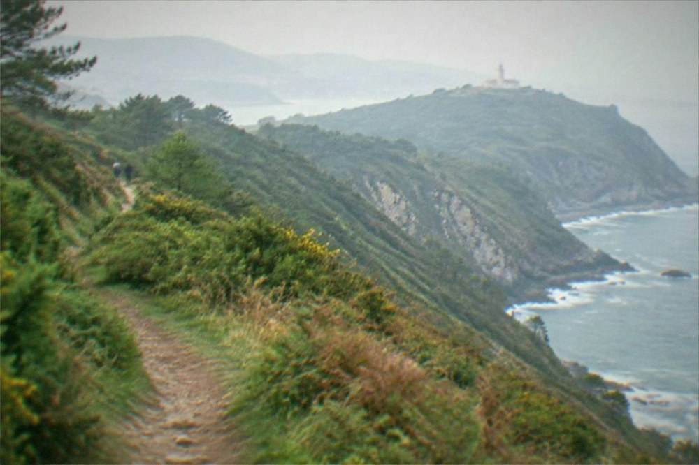 The coastal path on Monte Ulia above the sea with green hills and the lighthouse in the distance