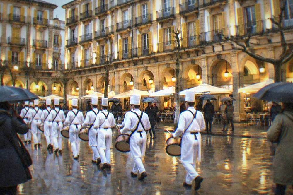 A drum company in white cook costumes marching past illuminated buildings at night