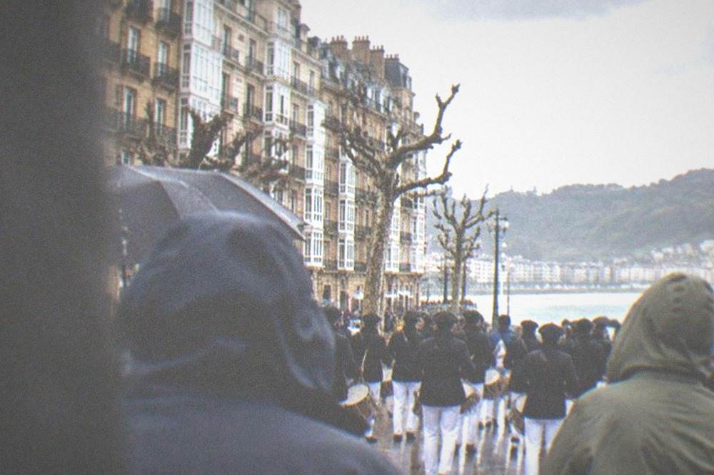 People in rain jackets watching a drum parade on a grey day beside a bay