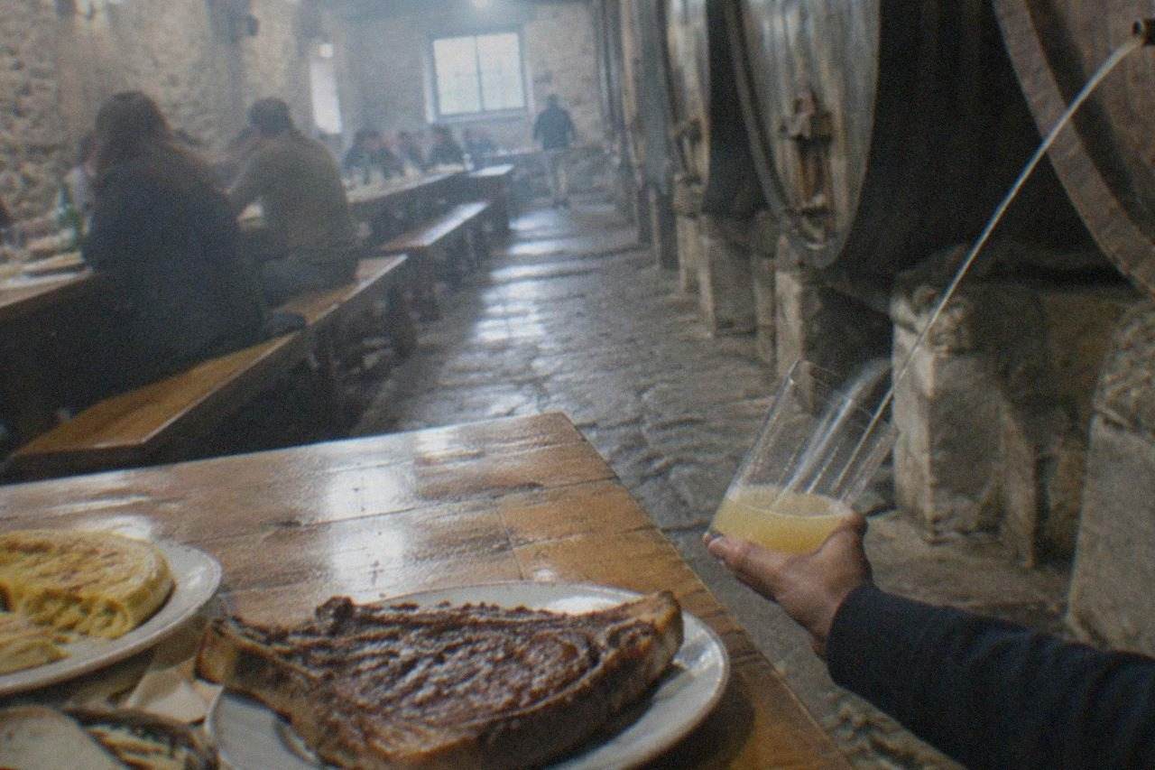 A thick bone-in rib steak resting on a grill over glowing embers in a Basque cider house
