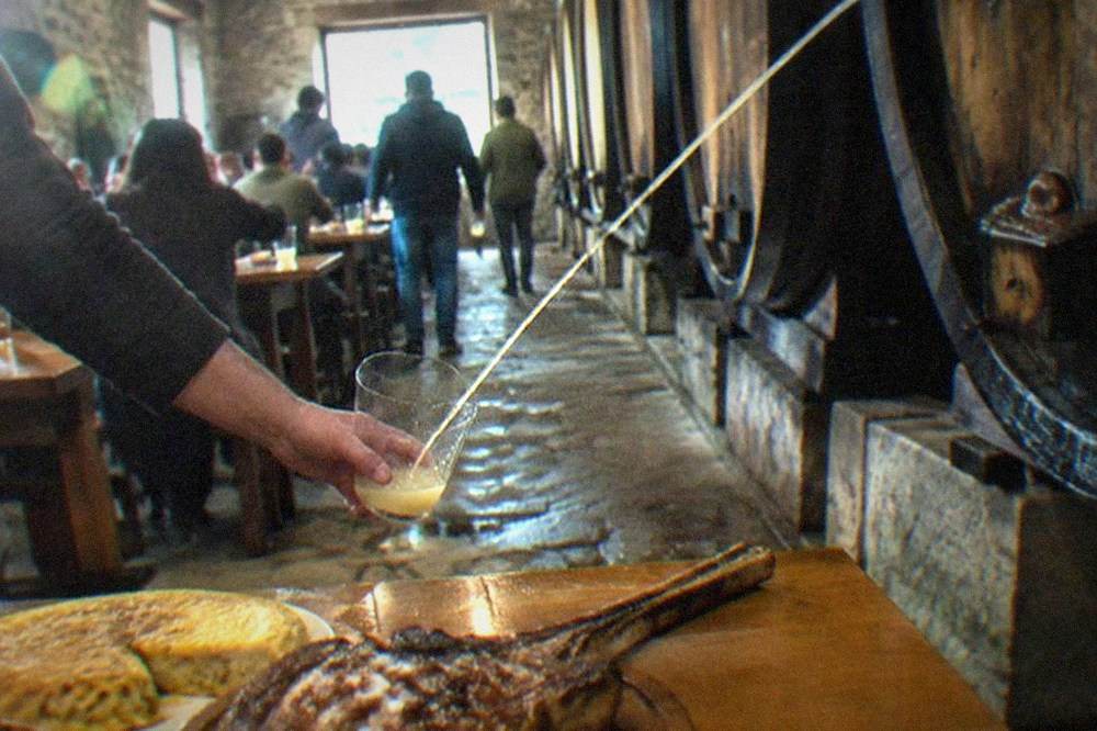Diners catching cider from a barrel spout in a crowded Basque cider house