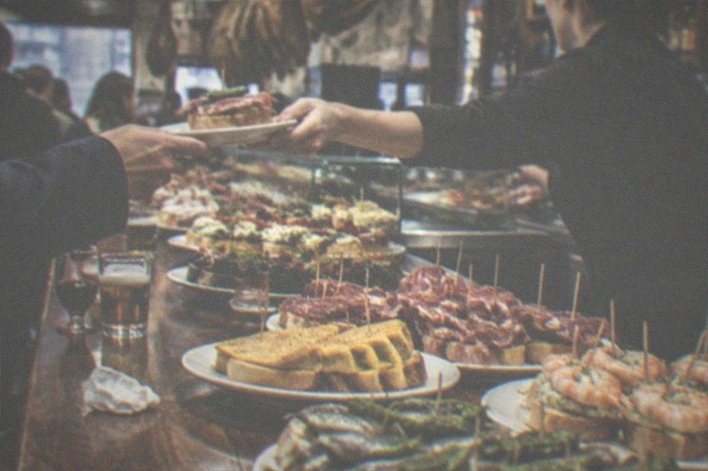 A bartender handing a small plate over a busy pintxo counter in the evening