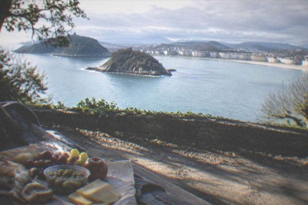A picnic of cheese, olives, bread and fruit on a bench overlooking La Concha bay