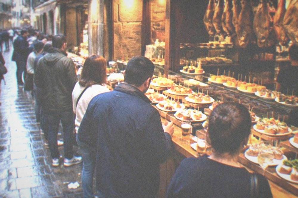 A narrow Old Town street in San Sebastián with pintxo bar fronts lit in the evening