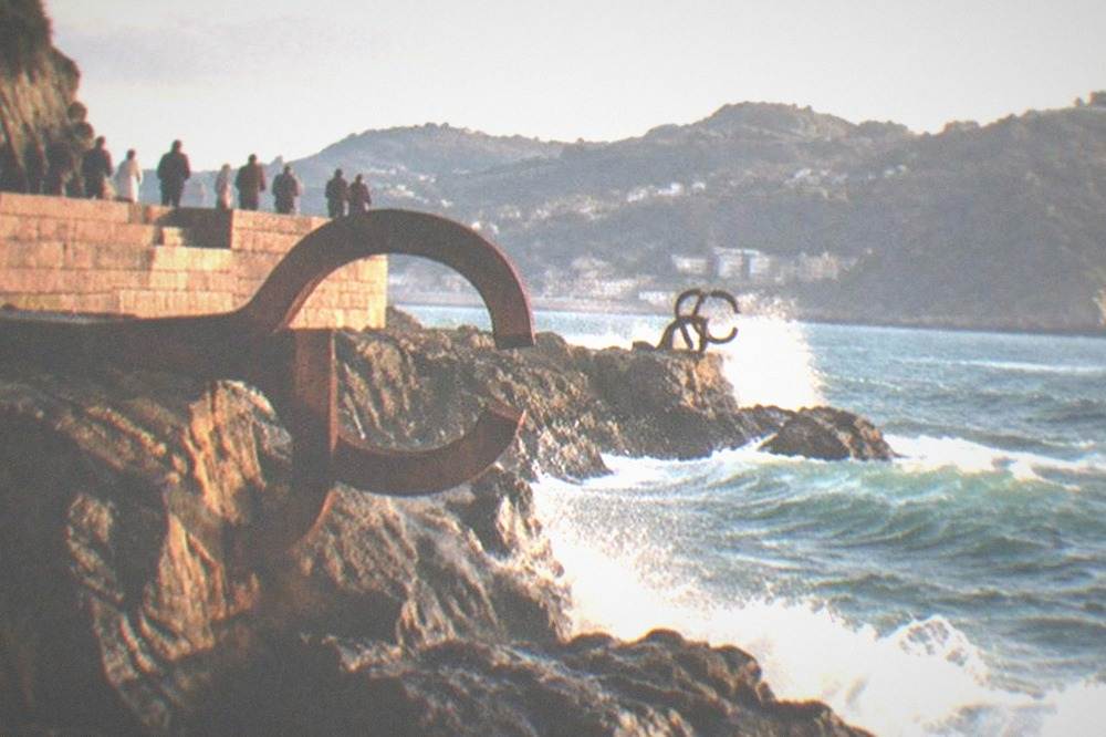 Steel sculptures of the Peine del Viento on rocks at the water's edge with waves breaking