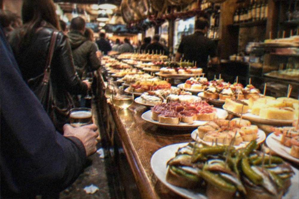 Crowds standing with drinks at a busy pintxo bar in the Parte Vieja at night