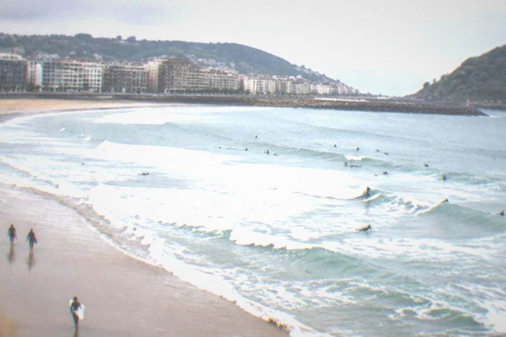 Zurriola beach with surfers in the water and the Gros neighbourhood's modern buildings behind the sand
